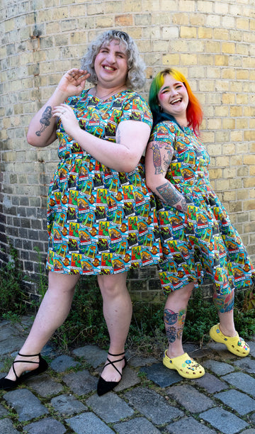 Two women standing and laughing together, wearing matching Run & Fly Dino Tarot stretch belted tea dress with pockets. The colourful dresses have an all over dinosaur tarot card print. One woman has curly blonde hair and the other has rainbow-coloured hair. Both have tattoos and are posing against a yellow brick wall. The dresses are from the ethical, slow fashion brand Run & Fly.