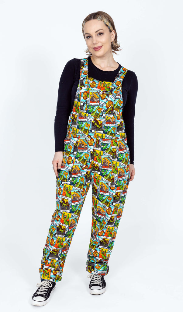 The Tarot Dinosaurs Stretch Twill Dungarees being worn by a femme model with short blonde hair with a long sleeve black top and black trainers. She is facing forward smiling with both hands by her side leaning back on one leg. The dungaree print has an aqua blue base with clouds with various scattered tarot cards with dinosaurs.