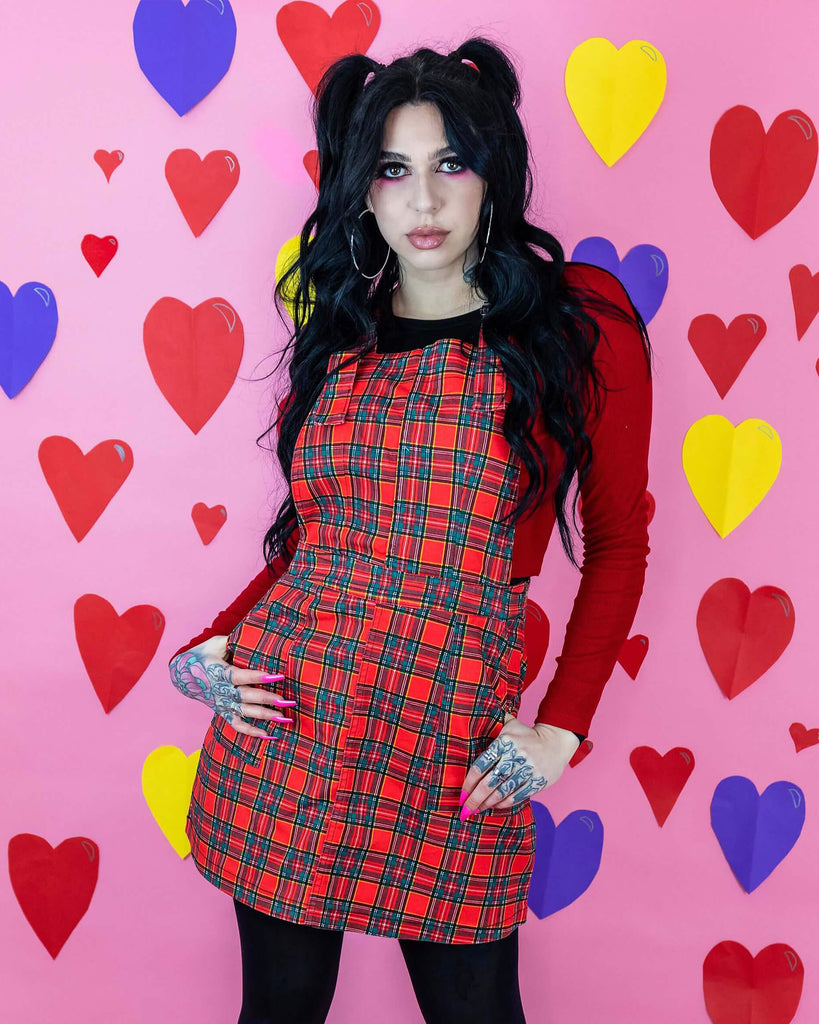 Rosie is stood in front of multicoloured hearts on a pink studio background wearing the red tartan pinafore dress with a long sleeve red tshirt, tights and black boots. They have long black hair and tattoos. They are facing the camera posing with both hands in the front pockets and looking to camera.