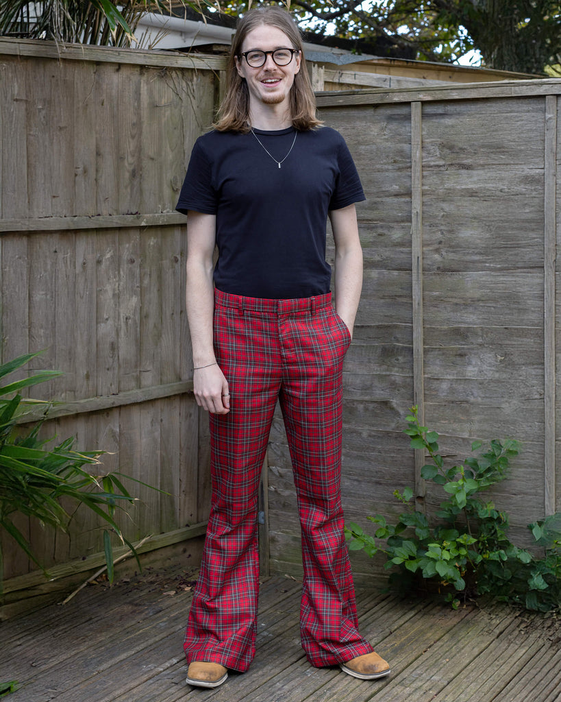 Jack is stood outside in front of a wooden fence wearing Retro Red Royal Stewart Tartan Plaid Bell Bottom Trousers with a black short sleeve t shirt and brown shoes. He is facing the camera and smiling with one hand in the trouser pocket.