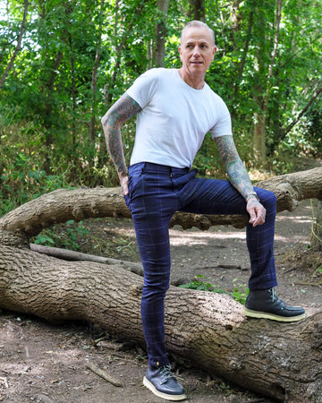 Jim is stood in a forest area wearing the retro mod stretch navy plaid tartan slim skinny fitting trousers with a white short sleeve tshirt and brown boat shoes. They are facing away from the camera with both hands in the side trouser pockets. 