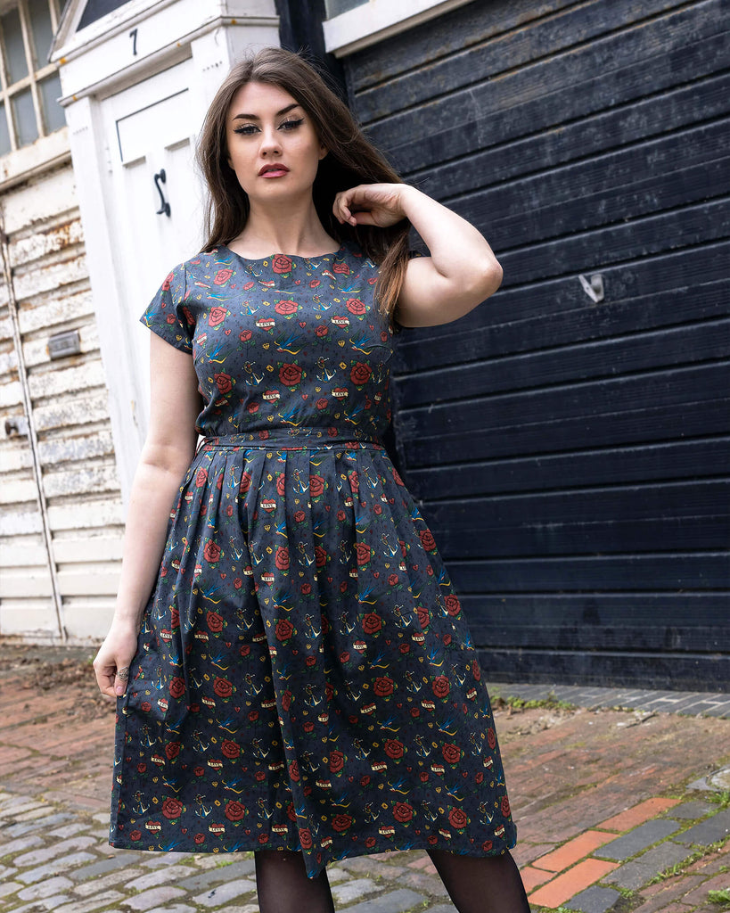 Charlotte is wearing the Run & Fly Retro Tattoo Stretch Belted Tea dress with pockets, grey with red roses, hearts and birds print, and waist belt. Model is looking at the camera, and standing in a cobbled street in front of garage doors with one hand in her hair.