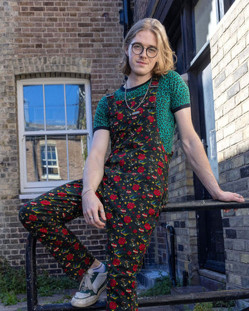 A long blonde hair male model with glasses wearing the run and fly retro old school tattoo stretch twill dungarees with the run & fly teal leopard short sleeve tshirt, green and white trainers and a silver chain necklace. They are sat on a metal rail leaning back with one hand propping them up and the other resting on their lap. The print features traditional tattoo designs such as roses and swallows.