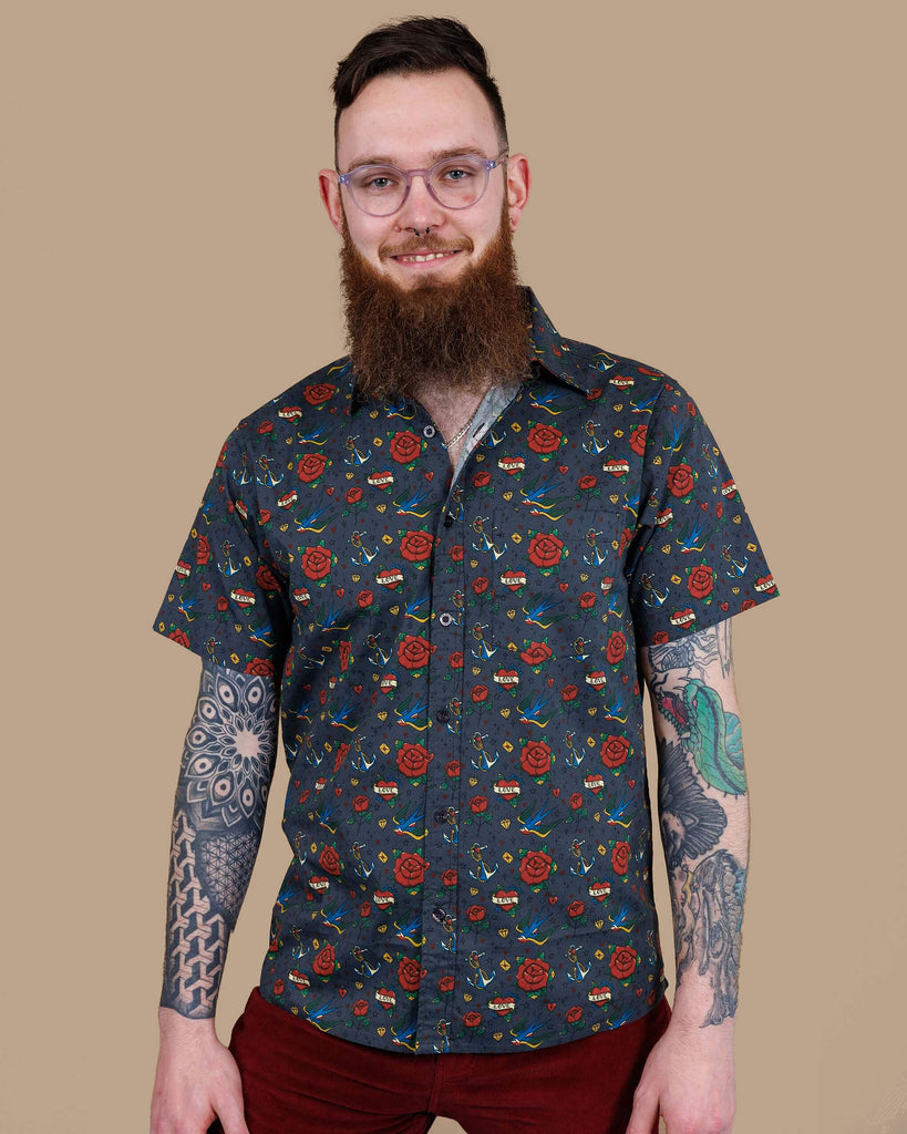 Dec, a white male model with a beard and glasses is wearing Retro Old School Tattoo Print Short Sleeve Shirt with Tawney Port Corduroy Bell Bottom Flares. The print has traditional tattoo style red roses, blue swallows, yellow envelopes, anchors and valentines love heart. Dec is stood in front of a beige backdrop at a photography studio in Hove facing the camera and smiling with his hands by his sides.