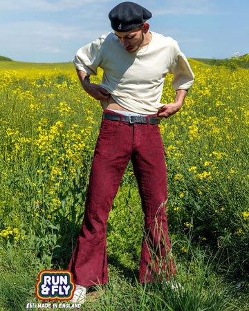 Zach is stood in a grassy flower field wearing the tawney port paisley corduroy bell bottom super flares with a short sleeve cream tshirt, black cap and white trainers. They are facing forward posing adjusting their tshirt whilst looking down to the left.