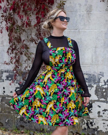 Karina a white female with blonde hair and sunglasses on is wearing a pinafore with yellow tigers and flowers on it with a black mesh top underneath smiling and stood outside against a white patchy wall