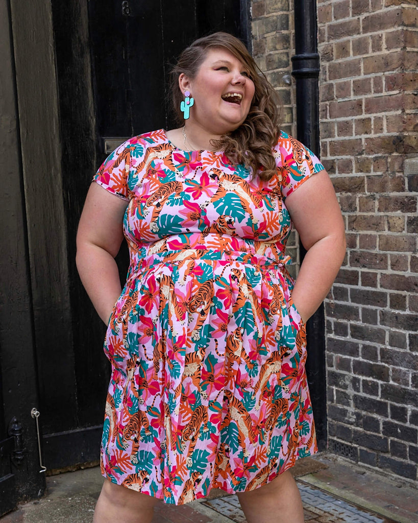 Natalie is wearing Tiger Lily Stretch Belted Tea Dress with Pockets with cactus shaped earrings and pink boots. The baby pink dress has an all over print of tigers with flowers and leaves. Natalie is stood outside in Hove facing the camera with her hands in the dress pockets and is smiling looking off to one side.