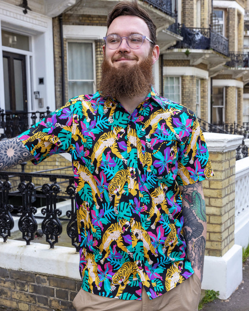 Tattooed model is wearing Black Tiger Lily Stretch Short Sleeve Shirt, paired with beige trousers. The collared shirt has a black background colour with all over orange tiger print and various foliage in pink, purple and green. Model is smiling and looking into the distance.