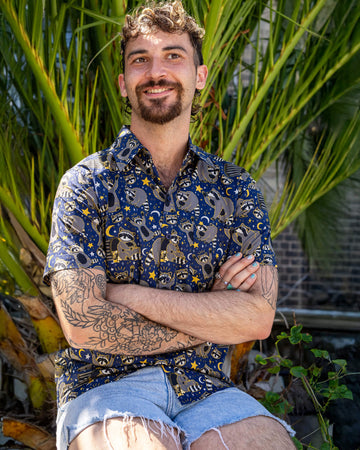 The Trash Pandas Short Sleeve Shirt worn by a masc non binary person with a curly brown mullet and denim shorts. They are sat outside on a white wall with green palm foliage behind them smiling looking right with their arms crossed. The shirt print features cheeky illustrated racoons, trash bins, rubbish, yellow moons and stars and white sparkles on a dark blue background.