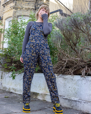 Model Julia wears Run & Fly dungarees featuring a whimsical print by Jen James Design with raccoons, moons, and stars. She stands outdoors, playfully covering her mouth and smiling, with bright yellow platform boots adding a bold touch to her outfit.