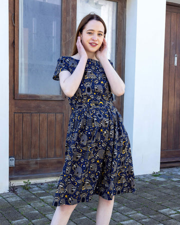 The Trash Pandas Stretch Belted Tea Dress with Pockets on a femme model with long brown hair and black heeled boots. She is stood posing outside in front of a wooden double door with her hands pushing back her hair smiling leaning on her right side. The print is a rich dark blue with grey racoons, trash bins, white and yellow sparkles, yellow stars and moons and small eyes.