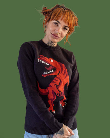 Amy is stood in front of a green background wearing the t rex dino-roar jumper with blue denim shorts. They have short ginger hair and tattoos. They are facing forward posing with their hands clasped in front of them whilst smiling to camera. Photo is cropped from the hips up.