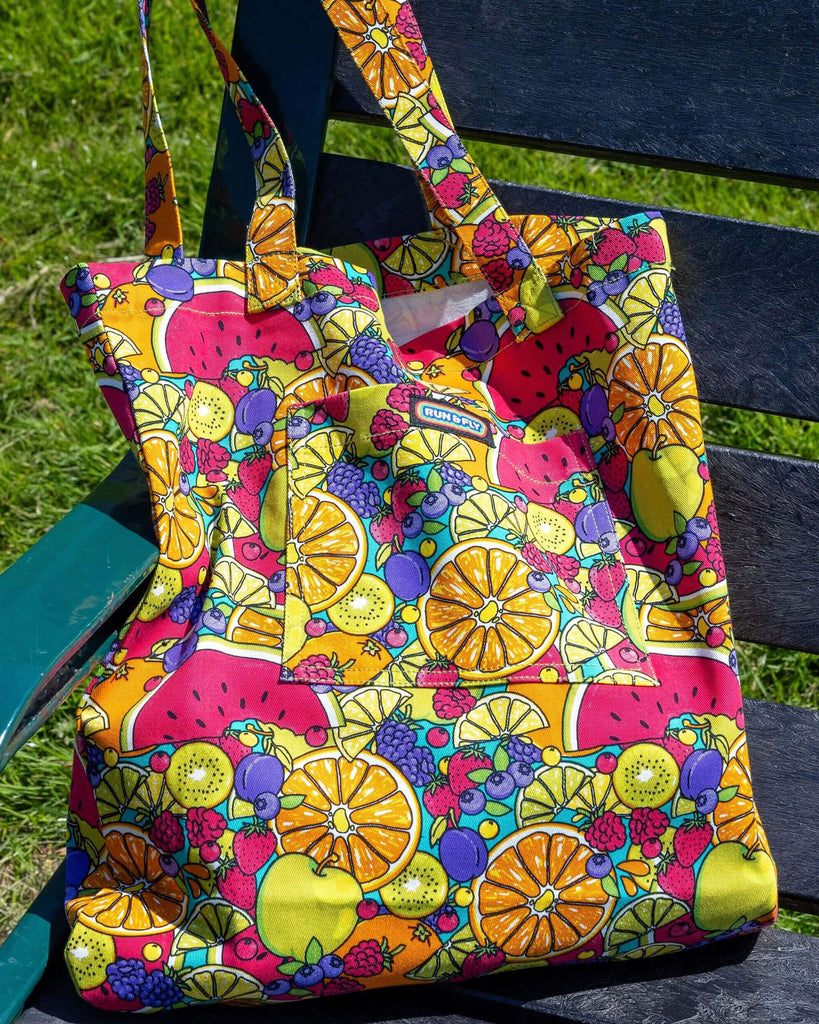 The Tutti Frutti Tote Bag laying on a dark wooden bench next to a grassy flowery field in the sunshine. The bright coloured tutti frutti tote bag has a front pocket with a rainbow run and fly label in the top middle. The tutti frutti print features berries, lemons, oranges, apples, watermelons, kiwis and plums all over on a blue background.