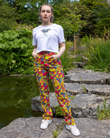 The Tutti Frutti Straight Leg High Waisted Jeans worn by a femme model with short blonde hair, a white butterfly crop top and white trainers. She is stood outside on a rock pathway dotted over a pond with green foliage and flowers surrounding. She is facing forward with both hands resting in the front pockets leaning more on one leg. The 90s style trousers feature various brighly coloured fruit slices on a turquoise blue base.