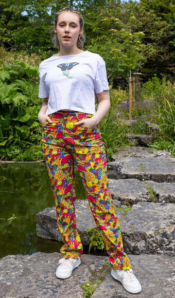 The Tutti Frutti Straight Leg High Waisted Jeans worn by a femme model with short blonde hair, a white butterfly crop top and white trainers. She is stood outside on a rock pathway dotted over a pond with green foliage and flowers surrounding. She is facing forward with both hands resting in the front pockets leaning more on one leg. The 90s style trousers feature various brighly coloured fruit slices on a turquoise blue base.