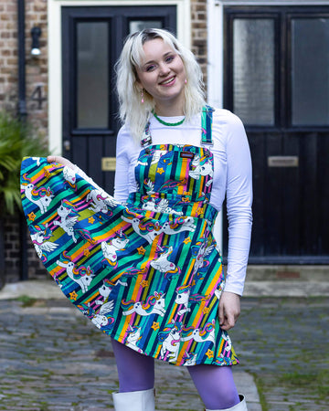 A blonde femme model wearing the unicorn stripes flared pinafore dress from run & fly with a long sleeve white top, purple tights and white platform boots. They are outside in a courtyard facing forward smiling whilst holding out the bottom of the pinafore. The slow fashion pinafore dress features rainbow stripes, kawaii cute pegasus', unicorns and stars with faces.