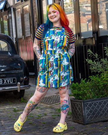 A model with rainbow-coloured hair and colourful makeup stood outside, wearing Unicorn Stripes Stretch Twill Pinafore Dress from ethical, slow fashion brand Run & Fly, paired with a rainbow striped t shirt.They are smiling and posing with their arms out by their sides, and visible tattoos on their arms and legs.