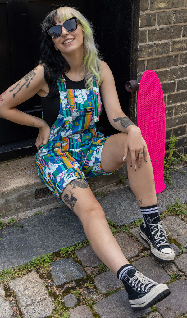 The woman sits on a step outside, smiling with her oversized sunglasses on. She is wearing a black tank top under her vibrant unicorn print short dungarees by Run & Fly, and black high-top Converse trainers. Her bright pink Penny skateboard is nearby.