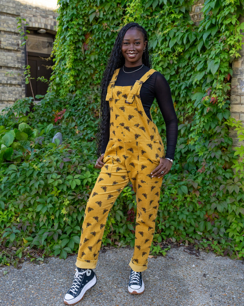 Model standing in mustard yellow bee dungarees from Run & Fly, smiling confidently in an outdoor setting filled with green ivy. Ethical slow fashion piece showcases black and orange embroidered bees and a comfortable fit, styled with black high-top trainers and a long-sleeve black mesh top.