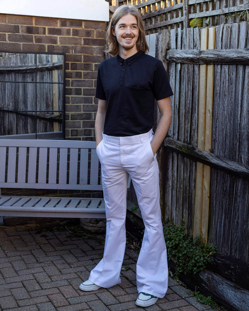 Jack is stood outside in a garden area wearing the Presley white cotton bell bottom trouser slacks with a short sleeve black shirt and white trainers. They have mid length blonde hair and are facing the camera posing with both hands in their front trouser pockets whilst looking off to the right smiling.