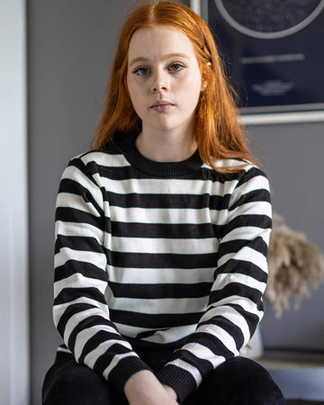 Sophie is sat against a grey background in front of a plant and has long red hair. They are wearing the White and Black Striped Jumper with dark jeans.