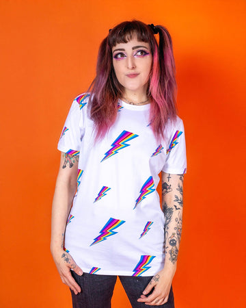 Sarah is stood wearing the white lightning bolt short sleeve tee with black stonewash denim flares. She has pink and brown hair in bunches with pink makeup and arm tattoos. She is facing towards the camera with both arms resting by her side and smiling looking up to the right. The tshirt is a white base colour with repeating rainbow lightning bolts varying in size all over.