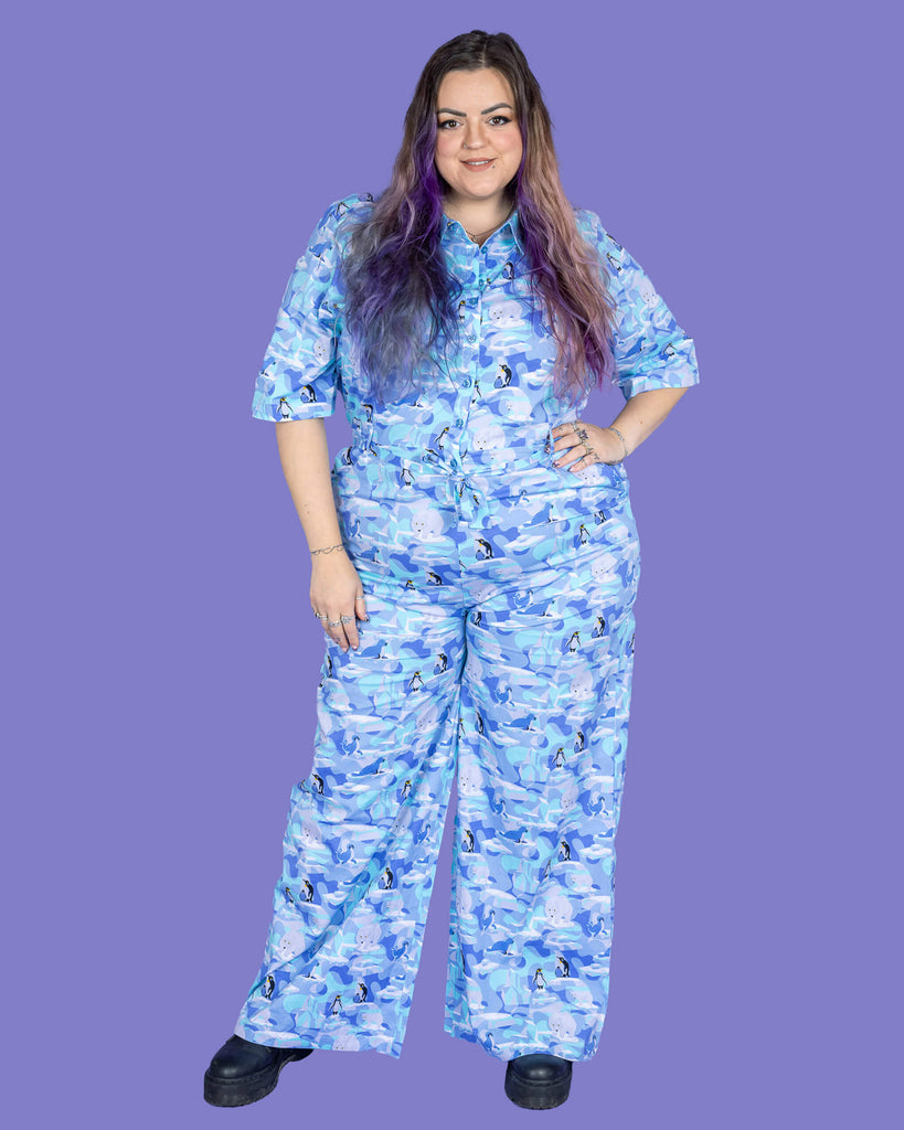 a model with purple hair wearing Winter Friends Stretch Jumpsuit paired with black boots. The jumpsuit is a light blue colour with ice burgs, seals, polar bears and penguins on. The model is smiling at the camera with one hand on her hip in front of a purple background.