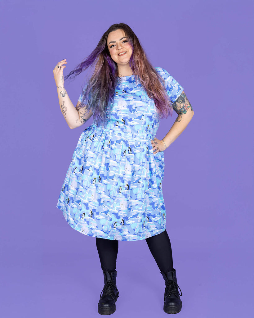 a model with purple hair wearing Winter Friends Stretch Belted Tea Dress with Pockets paired with black boots and tights. The jumpsuit is a light blue colour with ice burgs, seals, polar bears and penguins on. The model is smiling and flicking her hair in front of a purple background.