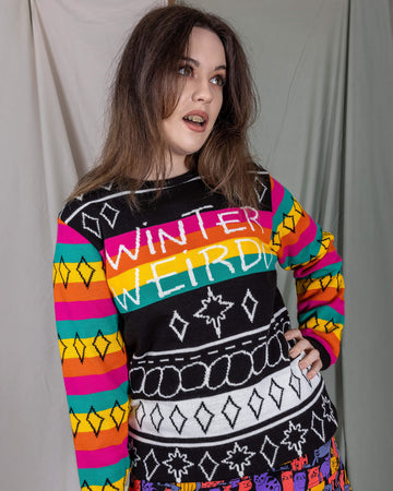 Alice is stood in front of a draped sheet background wearing the Run & Fly x Katie Abey Winter Weirdo Jumper with cats club pinafore underneath. She is facing forward with one hand on her hip, the other hand by her side whilst looking off to the right. The jumper has rainbow sleeves with black cuffs and sparkles, the chest is black and white christmas sparkles and patterns striped down it with 'Winter Weirdo' written in the centre with a rainbow behind the text to match the sleeves.