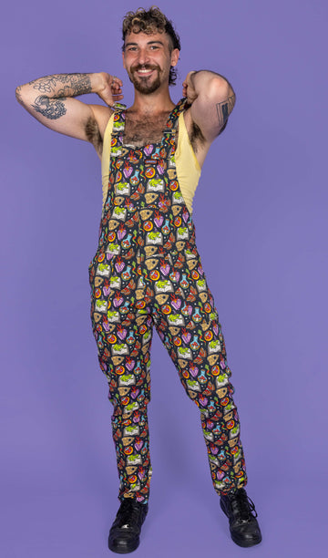 The Bewitched Stretch Twill Dungarees worn by a masc non binary person with a brown curly mullet, a yellow vest top and black trainers on a purple studio background. They are facing forward laughing with both hands reaching up to adjust the roller straps. The dark grey base dungarees feature colourful spell books, potion bottles, planchettes, snakes, frogs in witch hats, Death's-head hawkmoths, crystals and white sparkles all over.