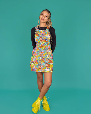 Milly, a femme model with blonde locs, is wearing Run & Fly x Katie Abey Weird & Wonderful Stretch Twill Pinafore Dress. The pinafore has cute bright Katie Abey illustrations all over. Milly is also wearing a long sleeved black top underneath the pinafore and bright green boots. Milly is smiling at the camera with her hands behind her back and one leg in front of the other. The background of the photo is forest green. 