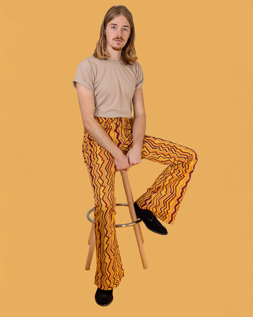 Jack is sat on a stool in front of a yellow studio background wearing the zig zag print bell bottom stretch super flares with a beige short sleeved tshirt and black boots. They are sat with one leg up on the stool foot rest to highlight the flares and both hands resting in the middle of their lap whilst looking to camera.